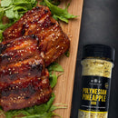 Two glazed ribs sprinkled with sesame seeds rest on a bed of arugula atop a wooden board, displayed beside a bottle of The Spice Lab Barbecue Seasoning Collection, all arranged on a dark surface.