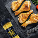Four seasoned chicken drumsticks rest on a slate plate alongside a bottle of The Spice Lab Barbecue Seasoning Collection’s Sweet + Tangy Mustard Rub, set on a dark surface with herbs and a cherry tomato—a mouthwatering addition from The Spice Lab for any barbecue seasoning collection.