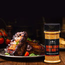 A plate of glazed ribs served with greens sits beside a bottle from The Spice Lab Barbecue Seasoning Collection by The Spice Lab, making it the perfect addition to any BBQ rubs lineup.