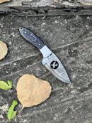 A Texan Knives FIXED BLADE POCKET KNIFE, a personalized Damascus steel hunting knife with a dark handle and patterned blade, rests on a weathered gray surface beside two brown leaves and one green leaf.