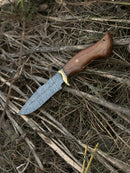 The Texan Knives Hunting Blade—a 10" Damascus steel knife with a rosewood handle, brass guard, and leather sheath—rests on a pile of dry branches and grass.