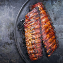 Two racks of grilled, glazed barbecue ribs on a dark, round tray with a rustic, textured gray background, seasoned to perfection with The Spice Lab Organic Granulated Onion by The Spice Lab—a versatile pantry staple that adds rich depth of flavor.