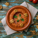A clay bowl of red dip flecked with The Spice Lab Aleppo Pepper Chili Flakes, garnished with walnuts, mint leaves, and garlic, sits on a beige napkin on a rustic blue wooden table with scattered walnuts and tomatoes.