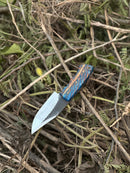 The Texan Knives Hunters Knife features a 9" D2 steel blade and an epoxy pine cone resin handle, shown resting on dry branches and green leaves outdoors; includes a leather sheath.