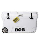 Introducing the Bob The Cooler Co. 75QT / 70.97L "BFF" Hard Cooler: a white cooler with black latches and extra thick, insulated walls for superior ice retention. Designed with heavy-duty wheels on one side, it features the Bob - The Cooler Co. lightning bolt logo on the front and a yellow tag attached to one latch reading “BOB THE COOLER CO.”.