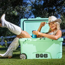 Lounging on the lawn, a woman in a white cowboy hat and boots relaxes inside the mint green Bob The Cooler Co. 75QT / 70.97L "BFF" Hard Cooler by Bob - The Cooler Co., smiling as she holds a chilled drink. Thanks to its extra thick insulated walls, her refreshment stays perfectly cold. In the background, trees and a white fence complete the scene.