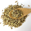 A wooden spoon filled with The Spice Lab Organic Fennel Seeds rests on a pile of more fennel seeds scattered across a white background, showcasing their mild licorice flavor—ideal for cooking and baking.