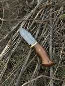 The Texan Knives Hunting Blade – a 10" Damascus steel knife with a rosewood handle, brass guard, and leather sheath – rests outdoors on a pile of dry sticks and grass.