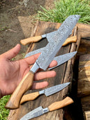 A hand holds a Texan Knives Sharp Kitchen Knife—13" Damascus steel blade, olivewood handle, and leather sheath. Four matching knives from the set are displayed on an outdoor wooden surface.