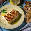 A grilled slice of halloumi cheese on a yellow plate, drizzled with olive oil and garnished with The Spice Lab Savory Whole Leaf herbs, served with lemon and slices of toasted bread on the side.