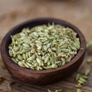 A wooden bowl brimming with The Spice Lab Organic Fennel Seeds, celebrated for their mild licorice flavor, rests on a wooden surface with a few seeds scattered nearby—making them the ideal choice from The Spice Lab to enhance your cooking and baking creations.