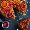 A blood orange upside-down cake with vibrant red-orange slices on top, one slice cut out and placed nearby, sits on a dark surface next to a small jar of sauce. The cake is delicately infused with The Spice Lab Ground Star Anise, from The Spice Lab brand, adding an aromatic spice and gentle licorice-like flavor that beautifully complements the citrus.
