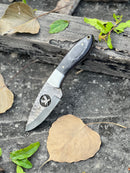 A Texan Knives FIXED BLADE POCKET KNIFE, a personalized Damascus steel hunting knife with a dark handle and bird-shaped blade cutout, rests on stone beside dry leaves, green leaves, and weathered wood.