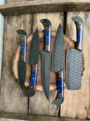 The Handmade Damascus Steel Chef Knife Set by Texan Knives features five textured black blades and dark wooden handles with blue resin accents—beautifully arranged, making it a perfect wedding or Valentine’s Day gift.