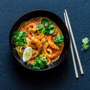 A bowl of noodle soup with shrimp, sliced green chili, fresh cilantro, and a lemon wedge sits on a dark surface. Chopsticks and a cilantro garnish are placed beside the bowl, highlighting the subtle aromas of The Spice Lab Organic Ground Coriander—an essential ingredient in Indian cooking spice blends from The Spice Lab.