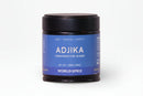 A small jar labeled World Spice Adjika by World Spice, featuring a blue background and a black lid. The label emphasizes its heat, vegetal and earthy notes, salt-free recipe, and signature fenugreek. Net weight is 1.98 oz (56g).