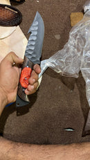 A person holds the Texan Knives Hunting Skinning Knife—a handmade 10" J2 steel blade with a Wangi/resin handle. Pieces of plastic, leather, and brown fabric are visible in the background.