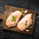 Two raw boneless chicken breasts from the Dude Food Gentleman’s Meat Box by Dude Food are arranged on a wooden cutting board, garnished with fresh basil leaves, garlic cloves, salt, and peppercorns, and set on a dark textured surface.