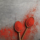 Two wooden spoons filled with The Spice Lab Organic Chili Powder Blend, a vibrant red seasoning from The Spice Lab and a staple in Mexican cuisine, rest on a gray textured surface with chili powder scattered around them.