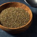 A wooden bowl filled with The Spice Lab Whole Celery Seeds sits on a dark textured surface, with a metal spoon partially visible in the upper right corner. These flavorful seeds from The Spice Lab are often used as savory seasoning or blended into pickling spices for added depth and aroma.