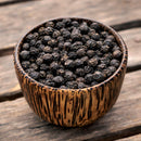 A small wooden bowl filled with The Spice Lab Special Extra Bold Indian Black Peppercorns sits on a rustic wooden surface.