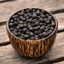 A small wooden bowl filled with The Spice Lab Organic Black Tellicherry Peppercorns Whole sits on a rustic wooden surface.