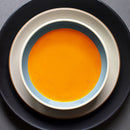 A bowl of vibrant orange soup, seasoned with The Spice Lab Nom Nom Paleo Magic Mushroom Powder from The Spice Lab for a rich umami flavor, rests on two stacked plates—one white and one black—as seen from above against a dark backdrop.