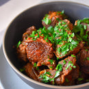 Chunks of braised beef in a dark sauce infused with The Spice Lab Nom Nom Paleo Magic Mushroom Powder, garnished with chopped fresh parsley, served in a dark bowl.