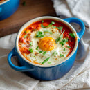 A baked egg rests atop a tomato-based sauce, enhanced with The Spice Lab Nom Nom Paleo Magic Mushroom Powder and garnished with fresh herbs, in a small blue casserole dish on a light cloth.