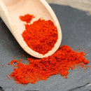 A wooden scoop with bright red paprika powder and a dash of The Spice Lab Cayenne Pepper X-Hot Ground spilling onto a dark slate surface.