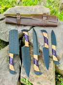 The Texan Knives Best Knife Block Set features a 13" forged stainless steel chef knife with an olivewood and epoxy resin handle, displayed with a leather sheath on rocks amid greenery for a striking presentation.