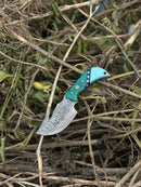 The Texan Knives Elite Custom Handmade Damascus Steel Guthook Skinning Knife features a distinctive resin handle and 7.5” patterned blade, shown resting on dry branches and leaves to highlight its premium craftsmanship and unique design.