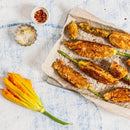 Battered and fried zucchini flowers on a parchment-lined tray, sprinkled with salt and The Spice Lab Organic Crushed Red Pepper Flakes. Loose yellow zucchini blossoms and small bowls of salt and The Spice Lab Organic Crushed Red Pepper Flakes sit beside the tray on a light blue background, adding an authentic Italian touch to this vibrant presentation.