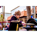 An elderly woman uses a WPPO 7" Round Turning Pizza Peel With Break Down Handle by WPPO LLC Direct to place a pizza into an outdoor oven, with people sitting at a table in the background and a tiled-roof building behind her.