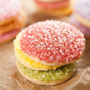 Three round sugar cookies stacked on a brown surface, each generously topped with The Spice Lab’s Sanding Con AA Sugar in sparkling colors—pink on top, yellow in the middle, and green on the bottom. Blurred cookies appear in the background, highlighting a charming touch of decorative baking.