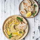 A plate of hummus drizzled with olive oil, garnished with The Spice Lab Organic Pine Nuts and fresh basil, served with pita bread slices. Another plate includes more pita, a lemon wedge, basil, and The Spice Lab Organic Pine Nuts—perfect as a salad topping or a versatile baking ingredient—all beautifully displayed on a white wooden surface.