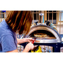 Using a WPPO 7" Round Turning Pizza Peel With Break Down Handle by WPPO LLC Direct, a person removes a cooked pizza from an outdoor oven, with stones and a fence visible in the background.