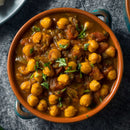 A bowl of chickpea curry garnished with chopped herbs, featuring whole chickpeas and tomatoes in a rich, spiced sauce infused with The Spice Lab Ancho Chile Powder by The Spice Lab, placed on a dark textured cloth.