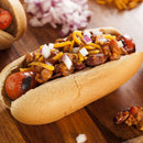 A close-up of a chili cheese hot dog topped with chopped onions, shredded cheddar cheese, and chili made with The Spice Lab Organic Chili Powder on a wooden board, with extra chopped onions and another hot dog in the background.