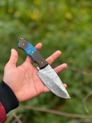 A hand holds the Texan Knives 10" Handmade Damascus Steel Hunting Knife, featuring a patterned blade and a resin handle in blue and dark brown, from their Gutting Knives collection, against a blurred green outdoor background.