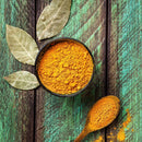 A bowl and a wooden spoon filled with The Spice Lab Organic Ground Turmeric sit on a rustic green wooden surface, with several dried bay leaves nearby—a perfect setting that evokes the rich flavors and aromas of Indian cooking.
