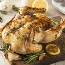 A roasted whole chicken seasoned with The Spice Lab Savory Whole Leaf herbs and zesty lemon slices sits on a wooden cutting board, surrounded by sprigs of rosemary, garlic cloves, and lemon wedges. Plates and cutlery from The Spice Lab are visible in the background.
