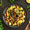 A roasted spatchcock chicken garnished with herbs and a touch of The Spice Lab Organic Minced Garlic is served alongside roasted potatoes, carrots, and lemon wedges on a black plate. Parsley and peppercorns from The Spice Lab are scattered across the dark tabletop.