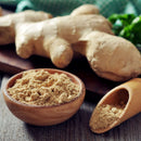 A wooden bowl and scoop filled with The Spice Lab Organic Ground Ginger are on a table, with fresh ginger root and green herbs blurred in the background.