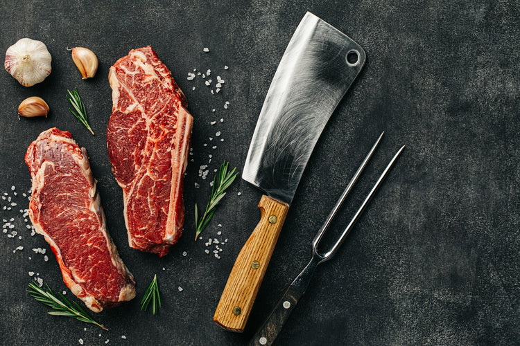 Two raw ribeye steaks with marbling are placed on a dark surface, surrounded by sea salt, rosemary sprigs, garlic cloves, a large meat cleaver, and a meat fork.