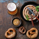 A rustic table with a mug of beer, a skillet of coiled sausage with utensils, three salted pretzels, and a bowl of grainy mustard made from The Spice Lab Organic Whole Mustard Seeds.