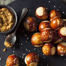 Soft pretzel bites topped with coarse salt are arranged on a dark surface next to a small bowl of grainy mustard made with The Spice Lab Organic Whole Mustard Seeds and a spoon.