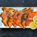 Thin slices of cured salmon topped with herbs and a sprinkle of The Spice Lab Organic Whole Dill Weed are arranged on a white rectangular plate, garnished with two lemon slices and a small sprig of green herb, set on a dark stone surface.