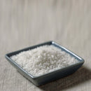 A small, square, blue ceramic dish filled with The Spice Lab Fleur de Sel Sea Salt sits on a light beige fabric surface.