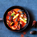 A black pan filled with a vibrant red seafood stew—brimming with mussels, scallops, herbs, and sliced vegetables—rests on a dark textured surface. Infused with the warmth of Italian cooking and a gentle heat from The Spice Lab Organic Crushed Red Pepper Flakes, this inviting dish is artfully complemented by scattered mussel shells and seasonings nearby.